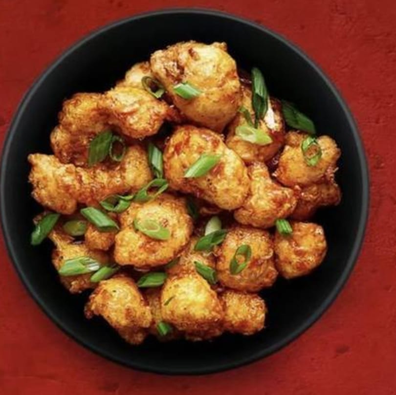 Golden fried cauliflower florets, garnished with fresh green onions, in a black bowl on a red background.