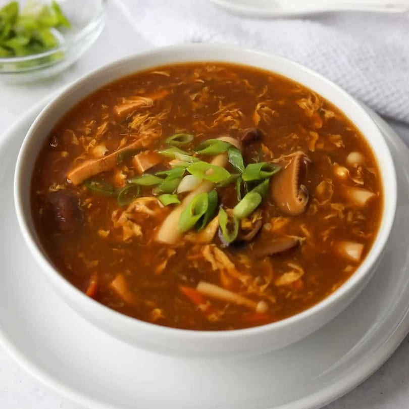 A bowl of hot and sour soup, garnished with green onions, mushrooms, and tofu.