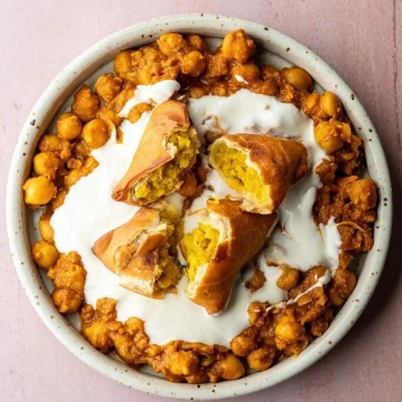 Indian samosas with yellow filling, chickpea curry, and white sauce in a speckled bowl.