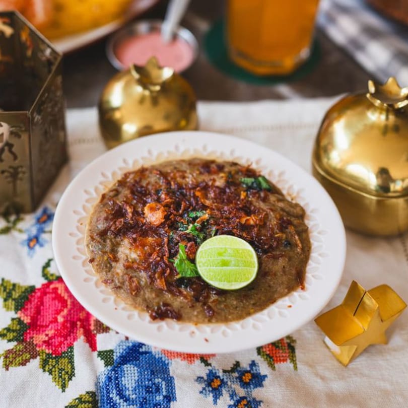 A plate of Indian Haleem, garnished with fried onions, mint, and a lime wedge, on a floral tablecloth.