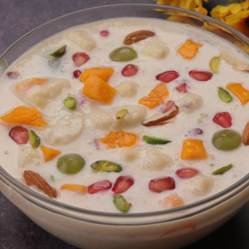Close-up of a glass bowl filled with Indian fruit custard, featuring colorful fruit pieces and nuts.