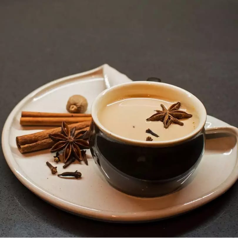 Indian chai tea in a black cup with star anise, cinnamon sticks, and cloves on a white plate.